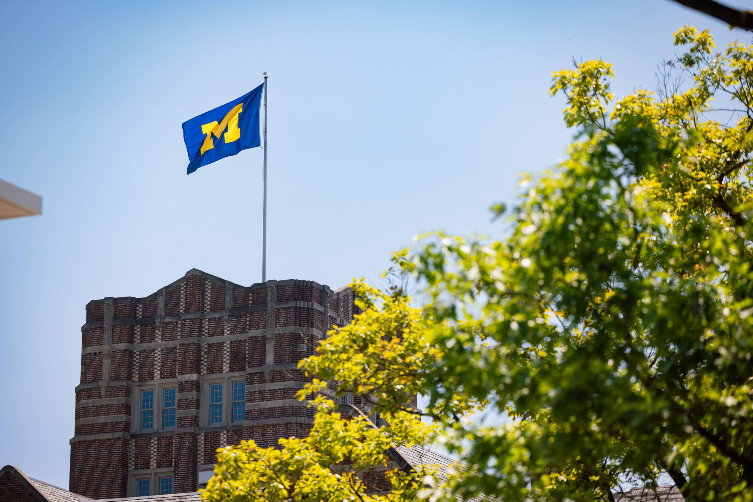 The flag on top of the Michigan Union waves against a clear blue sky on a sunny summer afternoon. A tree in the foreground obscures a bit of the Union’s dark brick tower.